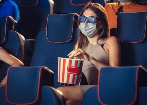 Cinema, movie theatre during quarantine. Coronavirus pandemic safety rules, social distance during movie watching. Men and women wearing protective face mask sitting in a rows of auditorium, eating popcorn.