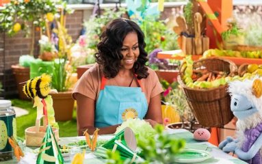 On a rooftop garden, Michelle Obama, wearing a blue apron, converses with a furry white and blue puppet with frozen waffle ears, a round, pink mochi puppet, and a bee puppet wearing a red tie around a table dressed with green decorations for a birthday party.