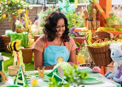 On a rooftop garden, Michelle Obama, wearing a blue apron, converses with a furry white and blue puppet with frozen waffle ears, a round, pink mochi puppet, and a bee puppet wearing a red tie around a table dressed with green decorations for a birthday party.